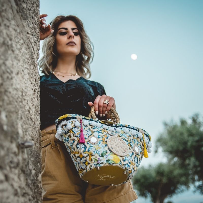 A person standing near a wall at dusk with a handbag featuring a colorful majolica and lemon patterned fabric, and stone mirrors.