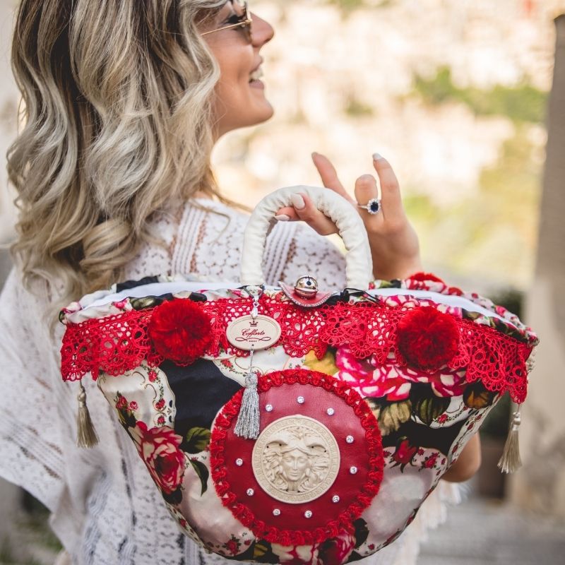 A close-up of a handbag with a floral pattern and red accents, featuring a bell closure and a central stone insert with a carved medusa woman surrounded by paillattes.